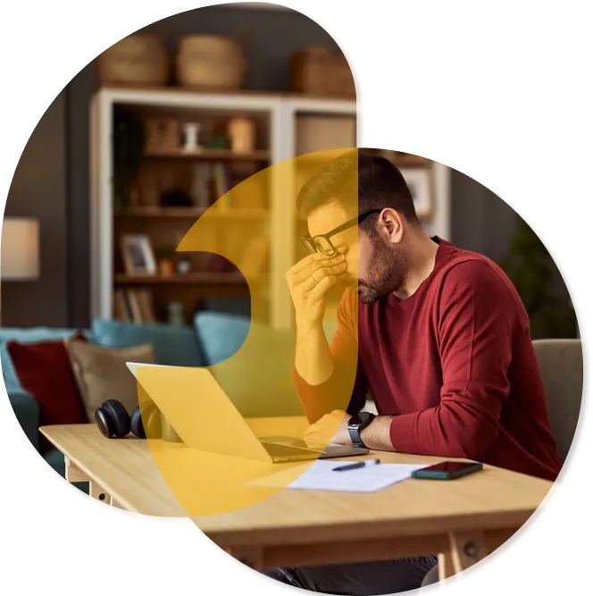 A frustrated man rubs his nose under his glasses while sitting at a desk with a laptop.