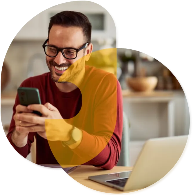 A man smiles while looking at his cellphone while sitting at a table with a laptop open.
